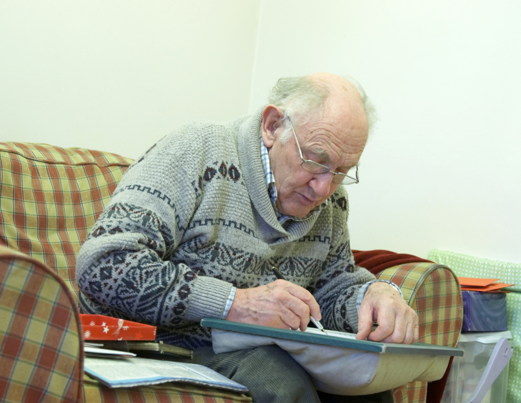 Older man in a grey jumper, writing a postcard in his home.